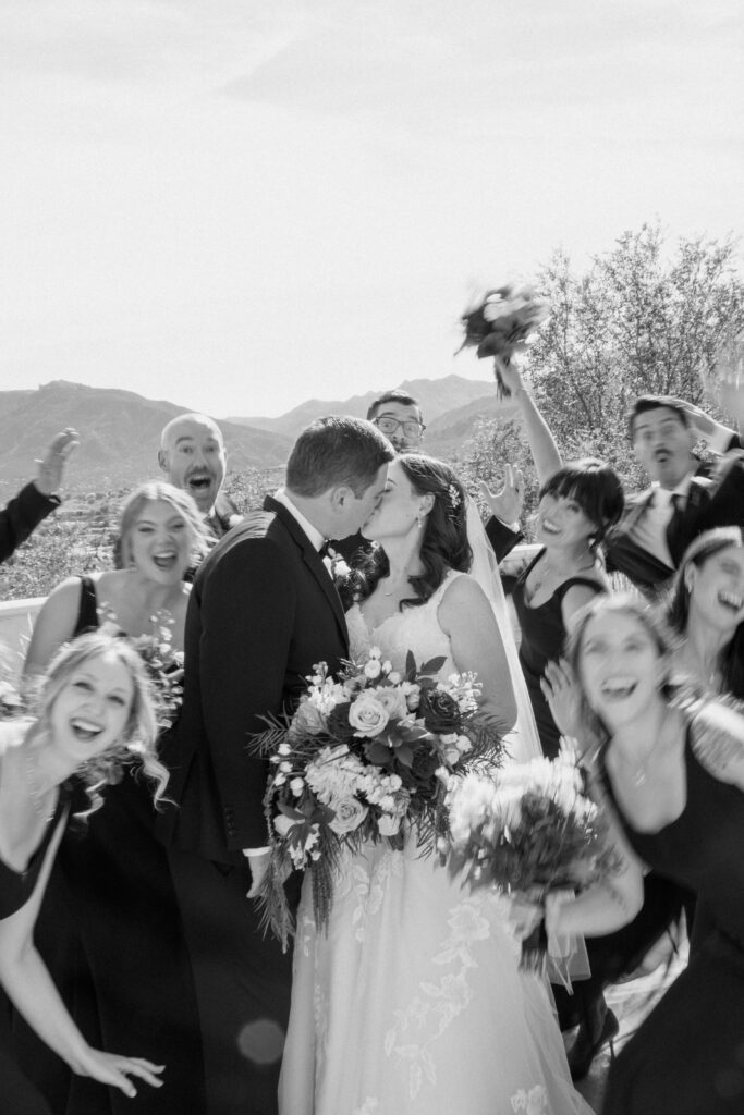 bride and groom kissing with wedding party cheering and mountain background
